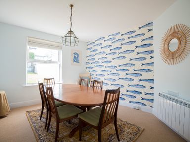 A dining room with a wooden table and six chairs a fish patterned wallpaper and a round wicker mirror at Tamarama House in Port Isaac