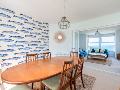 A dining room with a wooden table and chairs and fish wallpaper next to a living room with blue sofas and a large window at Tamarama House in Port Isaac