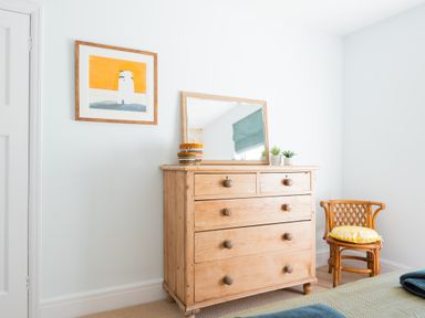 A wooden dresser with a mirror and small plants next to a wicker chair with a yellow cushion in a bedroom at Tamarama House in Port Isaac