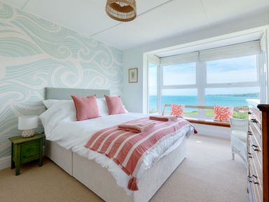 A bedroom with a bed, two pink pillows, pink striped blankets, a green bedside table with a lamp, a wooden bench with two coral patterned cushions by a large window overlooking the sea, a white wicker chair, and a wooden chest of drawers at Tamarama House in Port Isaac