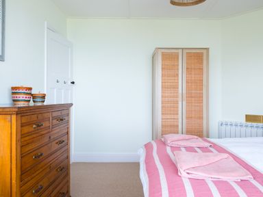 A bedroom with a wooden chest of drawers two baskets a wardrobe and a bed with pink and white striped bedding at Tamarama House in Port Isaac