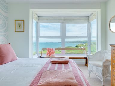 A bedroom with a bed facing large windows overlooking a coastal view with a bench and a wicker chair in the room at Tamarama House in Port Isaac