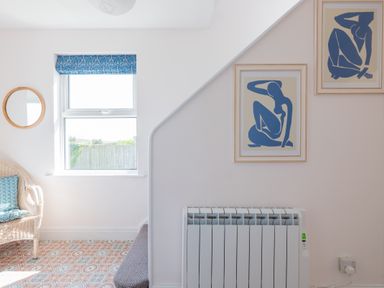 A room with a window a wicker chair with cushions a round wall mirror two framed art prints a radiator and patterned floor tiles at Tamarama House in Port Isaac