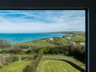A view of green hills and houses by the sea through a window at Tamarama House in Port Isaac