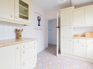 A kitchen with cream cabinets a tiled patterned floor a container of wooden utensils a bread box and a lobster picture on the wall at Tamarama House in Port Isaac