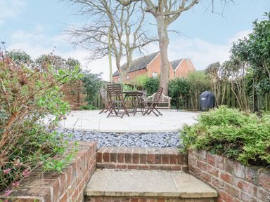 An outdoor garden area with a patio table and chairs surrounded by plants and trees at 9 Church Farm Rise in Aldeburgh