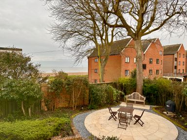 An outdoor seating area with a round paved patio table and chairs near trees and shrubs by red brick buildings at 9 Church Farm Rise in Aldeburgh