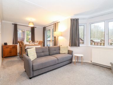 A living room with a grey sofa, side table, lamp, dining table with wicker chairs and windows at Hill Lodge Applethwaite 2 Limefitt Park near Windermere