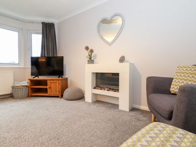A living room with a television on a wooden stand a white fireplace a heart-shaped mirror gray armchairs and a gray pouf at Hill Lodge in Applethwaite 2 Limefitt Park near Windermere