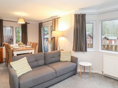 A living room with a gray sofa and two patterned pillows a small round table floor lamp and dining table with wicker chairs at Hill Lodge in Applethwaite 2 Limefitt Park near Windermere