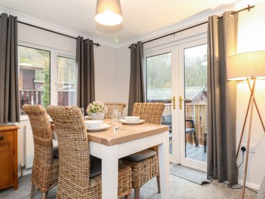 A dining area with a wooden table set with bowls and glasses four wicker chairs grey curtains and a glass door leading outside at Hill Lodge in Applethwaite 2 Limefitt Park near Windermere