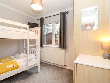 A bedroom with white bunk beds gray curtains a radiator and a wooden cupboard with a lamp at Hill Lodge in Applethwaite 2 Limefitt Park near Windermere