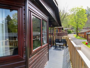 A wooden balcony with two chairs and a table overlooking a road and trees at Hill Lodge in Applethwaite 2 Limefitt Park near Windermere