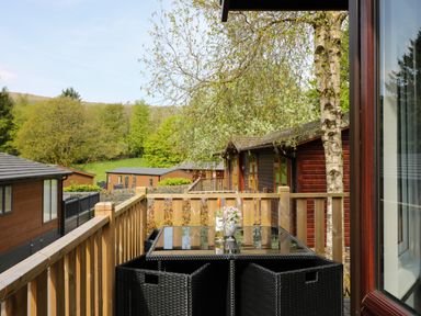 A balcony with a black glass table and four black wicker chairs surrounded by wooden railings overlooking brown wooden lodges and green trees at Hill Lodge Applethwaite 2 Limefitt Park near Windermere