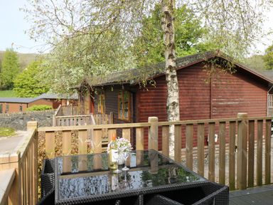 A wooden deck with a glass table and chairs overlooking wooden lodges and trees at Hill Lodge Applethwaite 2 Limefitt Park near Windermere