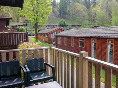 A balcony with two black chairs and wooden railings overlooking wooden cabins and a tree at Hill Lodge in Applethwaite 2 Limefitt Park near Windermere