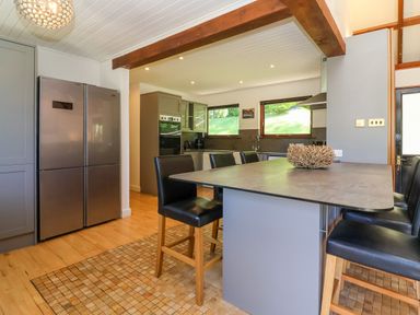 A kitchen with a large island and black stools a stainless steel refrigerator and windows showing green outside at Bay Tree Lodge 35 Watermouth near Berrynarbor