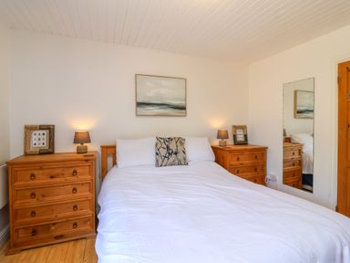 A bedroom with a double bed white bedding two wooden dressers with lamps and framed pictures a painting above the bed and a wall mirror at Bay Tree Lodge 35 Watermouth near Berrynarbor
