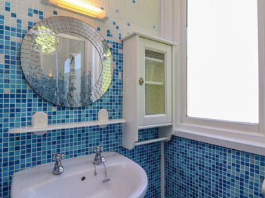 A bathroom sink with two taps under a round mirror and a white cabinet on a tiled blue wall at Bay Tree Lodge 35 Watermouth near Berrynarbor