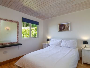 A bedroom with a double bed white bedding bedside tables with lamps a window with a blue blind and a wall mirror at Bay Tree Lodge 35 Watermouth near Berrynarbor