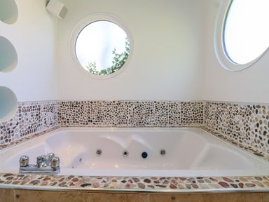 A bathroom with a corner bathtub surrounded by pebble tile walls and two round windows at Bay Tree Lodge 35 Watermouth near Berrynarbor