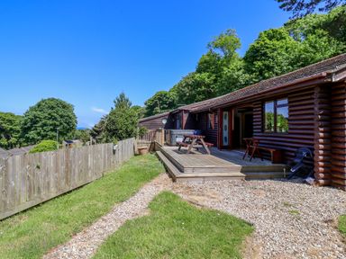 An outdoor wooden deck with picnic table and chairs beside a log cabin with trees and fence in the background at Bay Tree Lodge 35 Watermouth near Berrynarbor