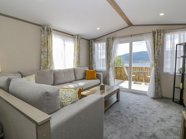 A living room with a gray sectional sofa and cushions by windows and glass doors opening to a balcony with chairs and a table at 11A Mansion View in Cove near Kilcreggan