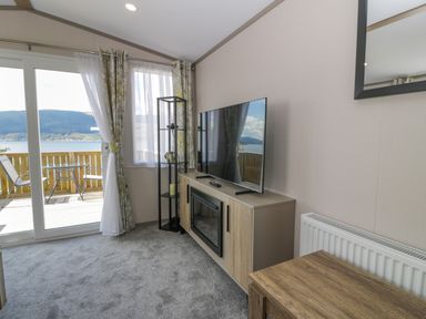 A living room with a television on a cabinet next to a glass door and window with outdoor seating visible at 11A Mansion View in Cove near Kilcreggan