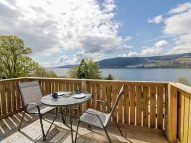 A wooden balcony with a glass table and two chairs overlooking water hills and trees at 11A Mansion View in Cove near Kilcreggan