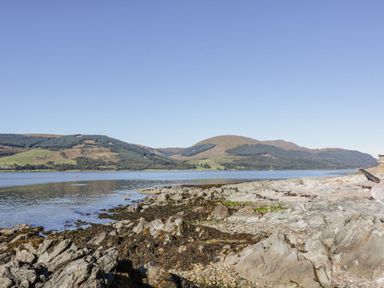 A rocky shoreline with calm water and hills in the background at 11A Mansion View in Cove near Kilcreggan