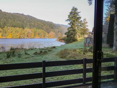 A view of a lake and forest from behind a wooden fence and glass door at 27 Invergarry Lodges in South Laggan near Invergarry