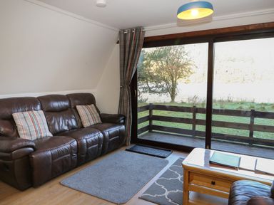 A living room with a brown leather sofa, two plaid cushions, a wooden coffee table with books, rugs on the floor, glass sliding doors, and a view of a wooden balcony and trees at 27 Invergarry Lodges in South Laggan near Invergarry