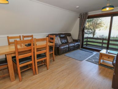 A living room with wooden dining table and chairs a leather sofa and glass doors overlooking a garden at 27 Invergarry Lodges in South Laggan near Invergarry