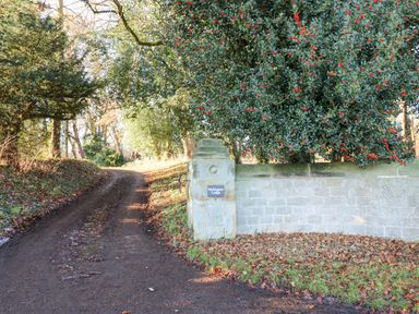 A dirt driveway beside a stone wall with a sign reading Wellington Lodge under trees with red berries at Wellington Lodge Cottage in Middleton Tyas