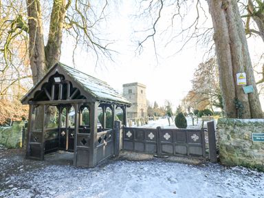 A wooden lychgate entrance gate leading to a churchyard with gravestones and a stone church tower visible at Wellington Lodge Cottage in Middleton Tyas