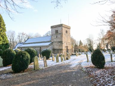 A graveyard with headstones and trimmed bushes next to a stone church with a square tower under a clear sky at Wellington Lodge Cottage in Middleton Tyas