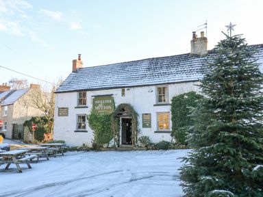 A snow-covered outdoor area with picnic tables in front of a white building with green signs and a decorated Christmas tree at Shoulder of Mutton in Middleton Tyas