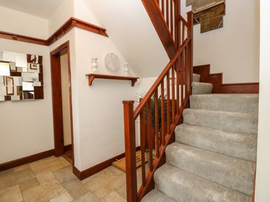 A stairway with carpeted steps and wooden railing next to a tiled floor hallway with a wall shelf and decorative items at Winding Wheel Cottage in Cambois near Ashington