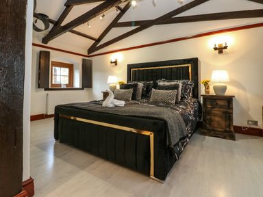 A bedroom with a large bed with dark upholstery and gold trim bedside tables with lamps and flowers exposed ceiling beams at Winding Wheel Cottage in Cambois near Ashington