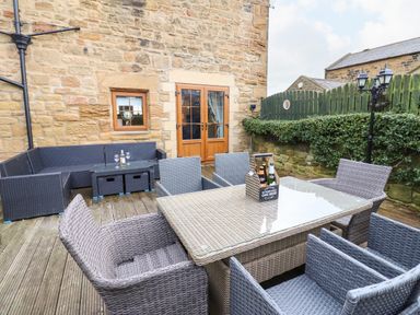 An outdoor patio with a wicker dining table and chairs and a wicker sofa set against a stone building at Winding Wheel Cottage in Cambois near Ashington