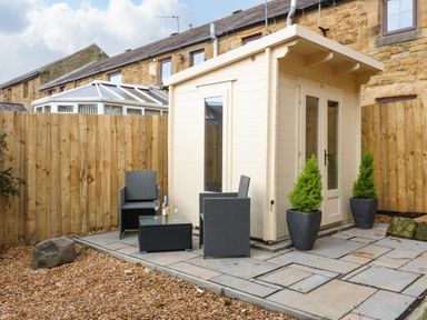 A small wooden shed with glass doors and windows on a stone patio with two black wicker chairs a table with a bottle and glasses and two potted plants at Winding Wheel Cottage in Cambois near Ashington