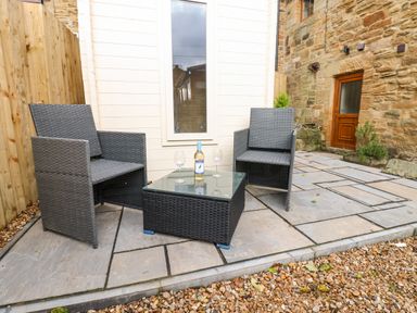 An outdoor seating area with two grey chairs a square glass-top table a bottle and two wine glasses on the table and stone and gravel ground at Winding Wheel Cottage in Cambois near Ashington