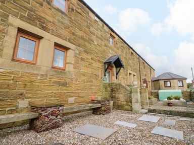 The exterior of a stone building with wooden framed windows and a gravel courtyard with stepping stones at Winding Wheel Cottage in Cambois near Ashington