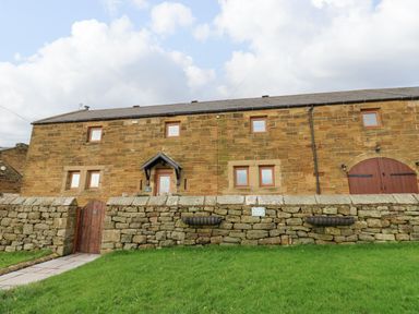 Exterior view of a stone building with wooden doors and windows behind a stone wall with a wooden gate and green lawn at Winding Wheel Cottage in Cambois near Ashington