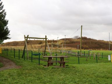 A playground with swings and wooden benches surrounded by a green fence in a grassy area at Winding Wheel Cottage in Cambois near Ashington