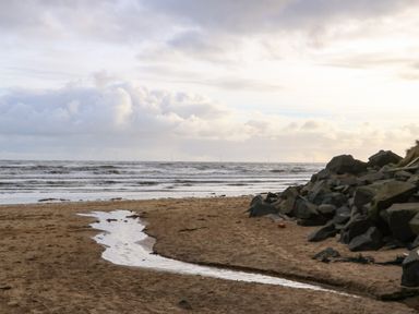 A beach with wet sand and a small stream flowing towards the sea with rocks on the right side at Winding Wheel Cottage in Cambois near Ashington