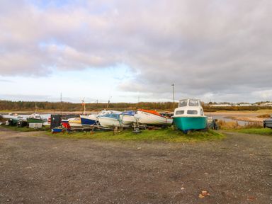 A group of boats parked on grass near a river with trees and buildings in the background at Winding Wheel Cottage in Cambois near Ashington