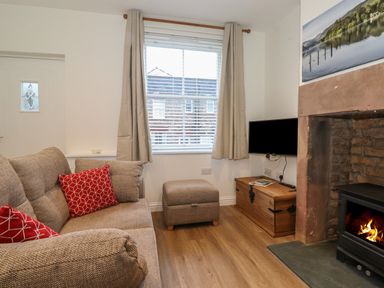 A living room with a sofa and red cushions a window with curtains a wood storage chest a mounted television and a fireplace at Jack's Cottage in Keswick
