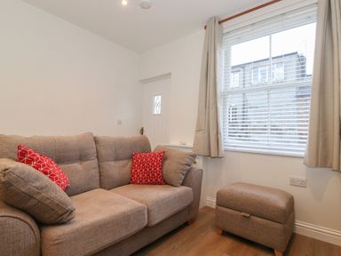 A small living room with a beige sofa with red cushions and a matching ottoman near a window with blinds and curtains at Jacks Cottage in Keswick