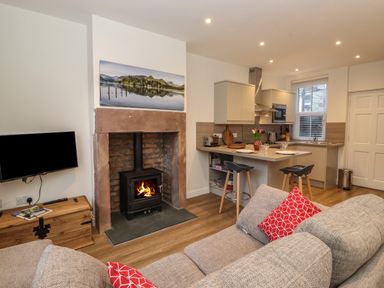 A living room with a fireplace a wall-mounted television a kitchen island with two stools and a sofa with red cushions at Jack's Cottage in Keswick